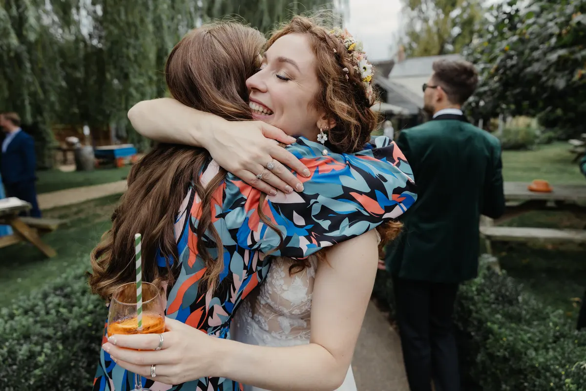 Bride in a flower crown smiling as a guest in a patterned dress hugs her in a pub garden