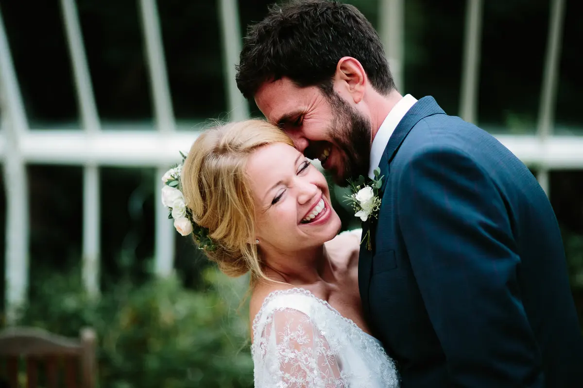 Groom nuzzling the laughing bride, who wears white flowers in her hair, by a glasshouse