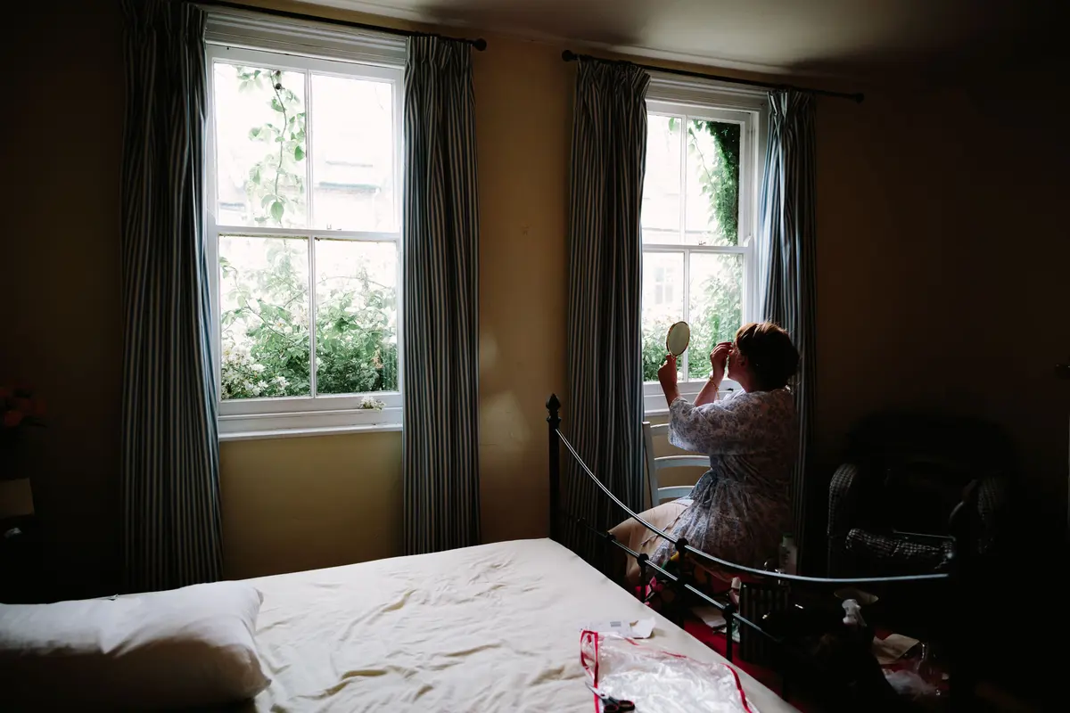 Bride in a robe doing her make-up with a hand mirror by a bedroom window