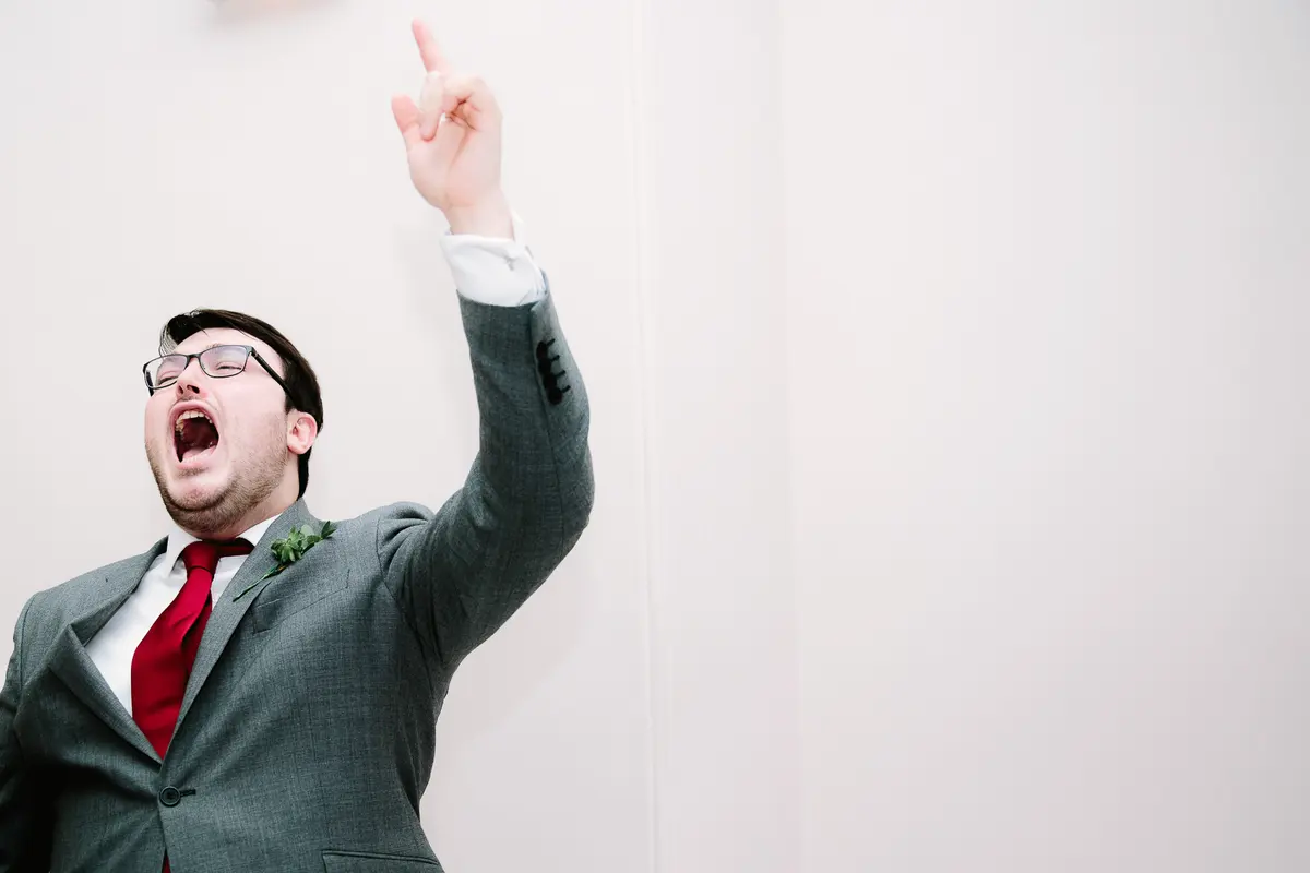 Man in a grey suit and red tie pointing upwards and shouting mid speech