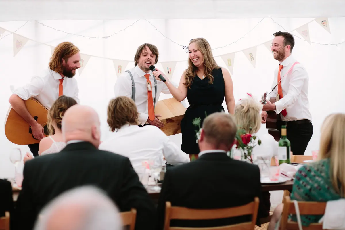Guitarists and a singer performing a song to the tables in a marquee hung with bunting
