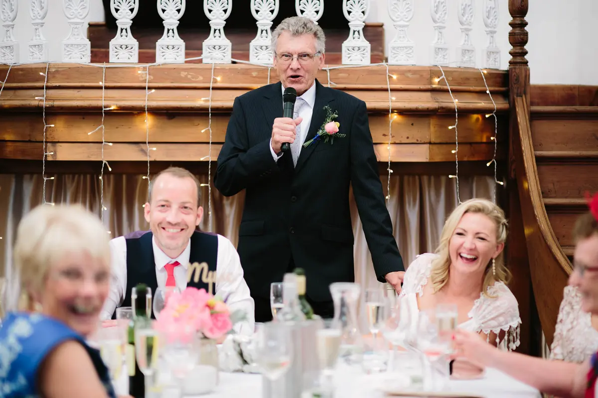 Older man giving a speech as the bride and groom laugh at the top table