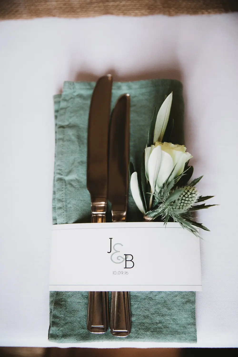 Place setting with a knife, fork, green linen napkin and a white rose tucked into a monogrammed band