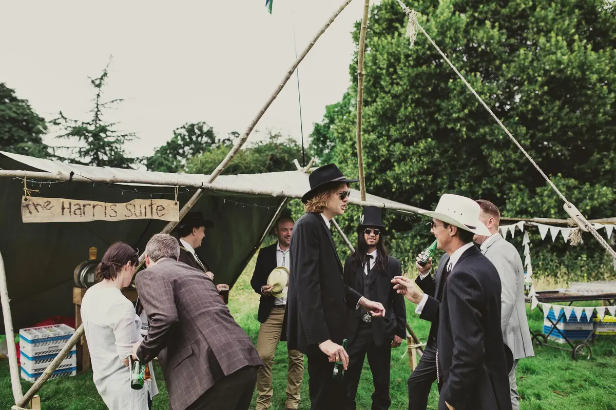 Guests in hats and suits chatting with drinks by a canvas bar tent hung with bunting