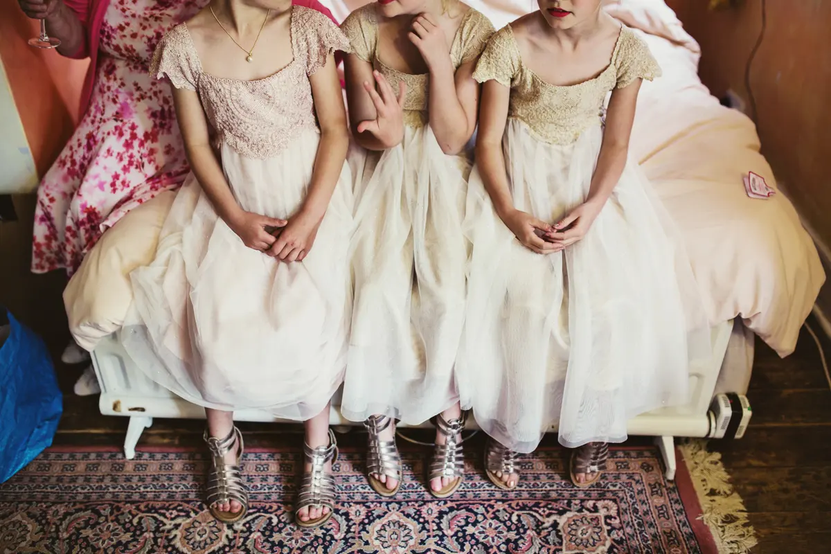 Three flower girls in lace and tulle dresses and silver sandals sitting in a row on a bed