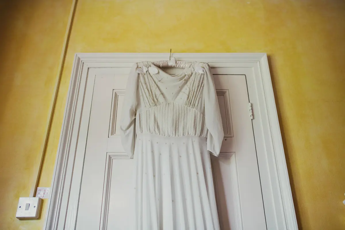 Vintage-style wedding dress with long sleeves hanging on a white door against a yellow wall