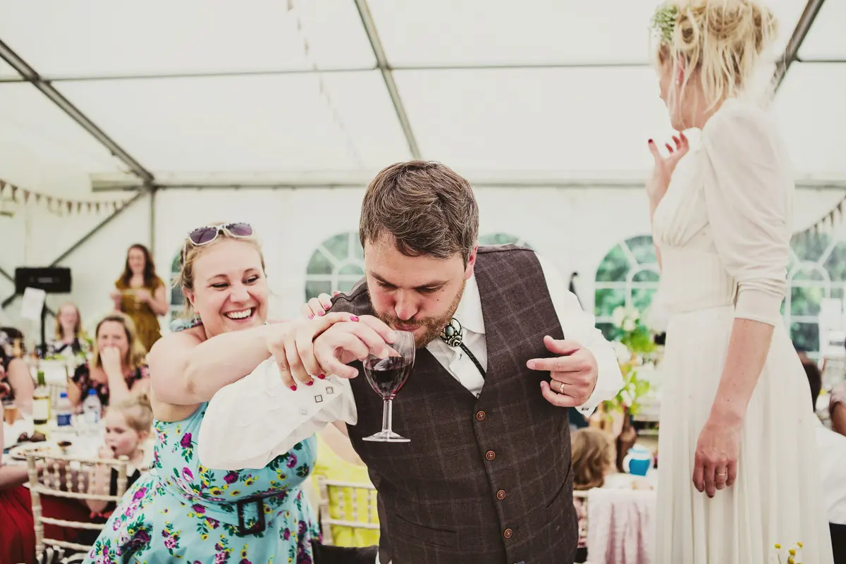 Laughing guest linking arms with the groom as he drinks red wine while the bride stands on a chair