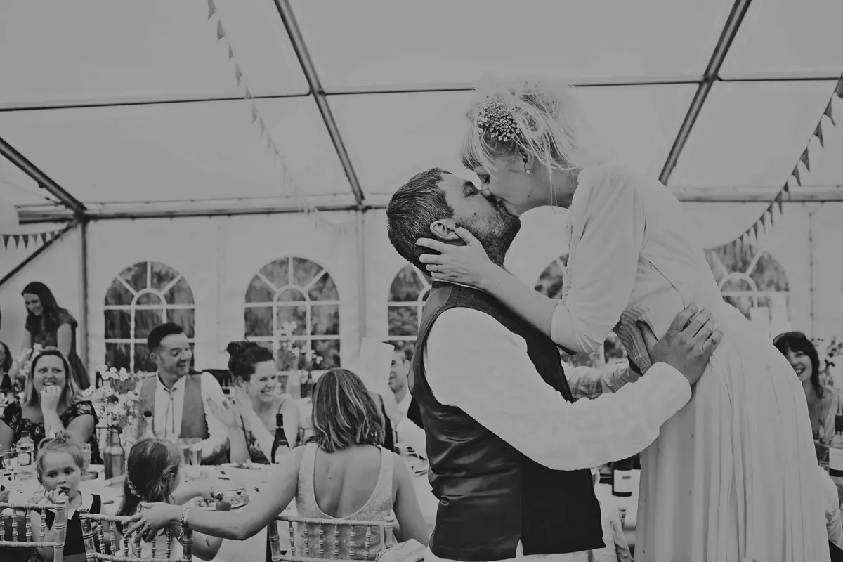 Black and white shot of a bride holding the groom's face and kissing him in a marquee as guests cheer