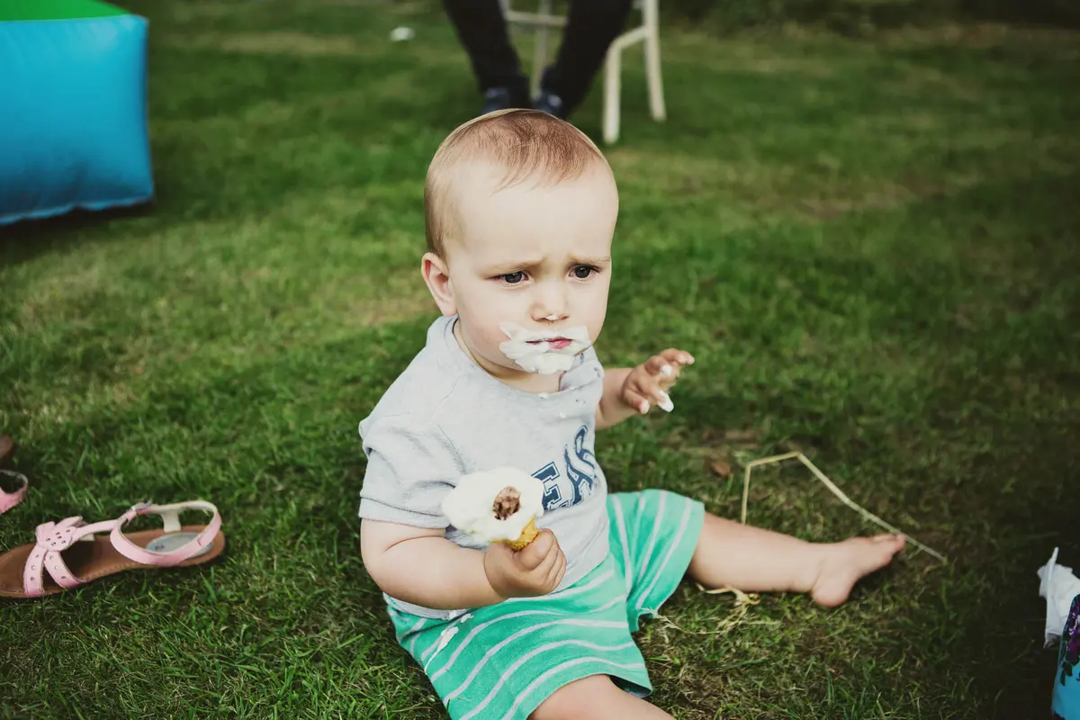 Toddler sitting on the grass with a cream moustache, holding a half-eaten cake