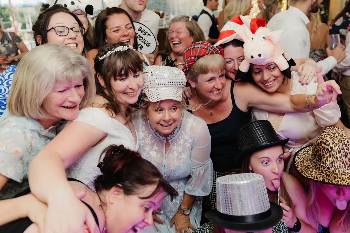 Bride hugging a crowd of laughing guests wearing sparkly party hats and photo booth props