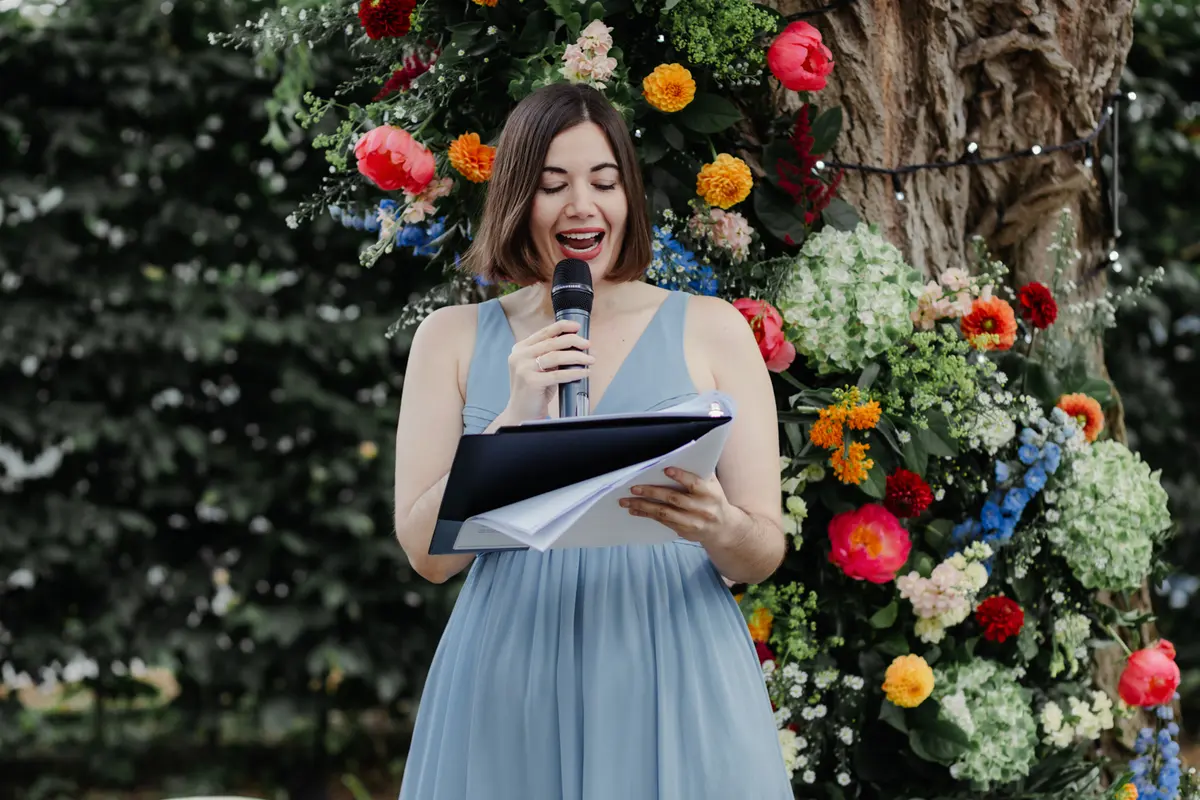 Woman in a blue dress giving a reading in front of a colourful flower arch at the Perch