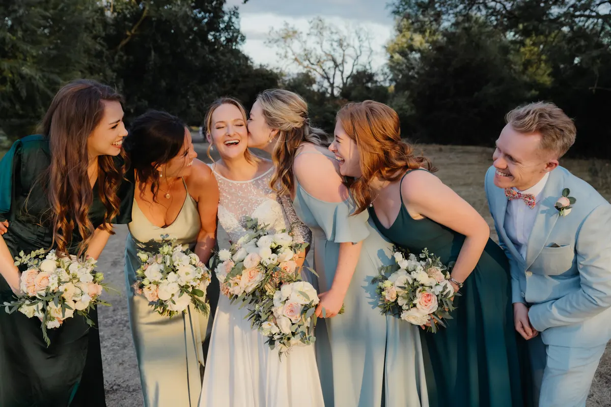 Bride laughing with her bridesmaids in green and a friend in a pale blue suit at golden hour