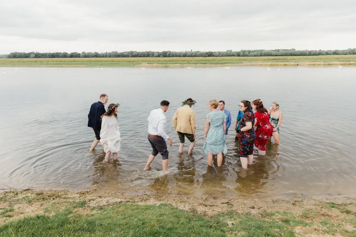 Bride, groom and guests wading barefoot into the shallow water at Port Meadow