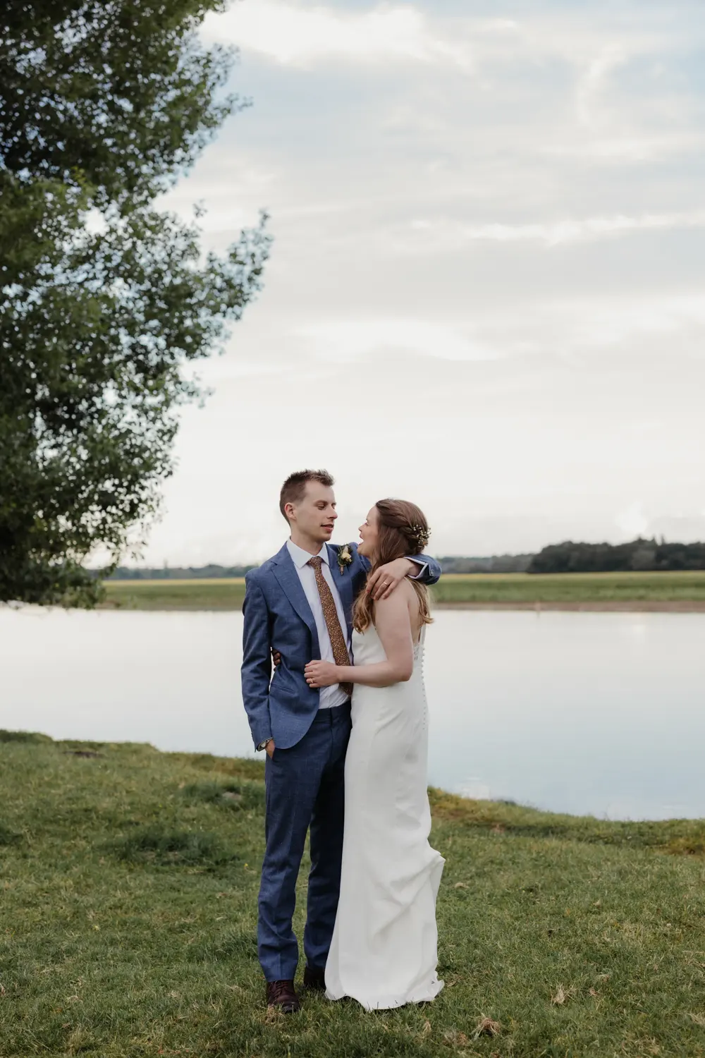 Bride and groom embracing on the grassy bank of the river at Port Meadow