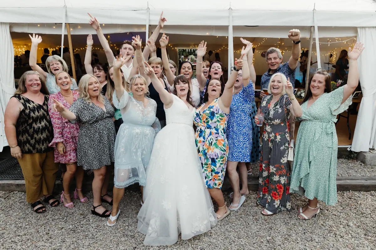 Bride with arms raised among cheering female guests outside the marquee