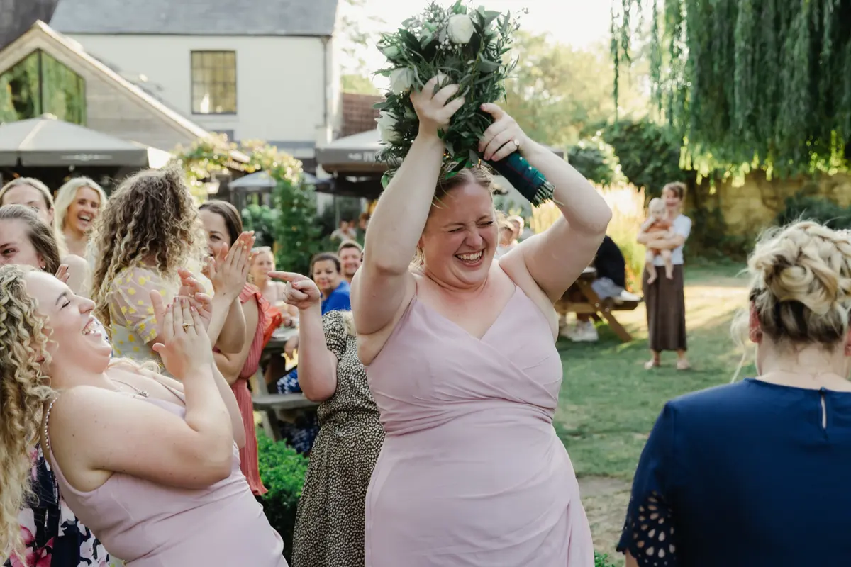 Bridesmaid in pink catching the bouquet as guests cheer in the garden at the Perch