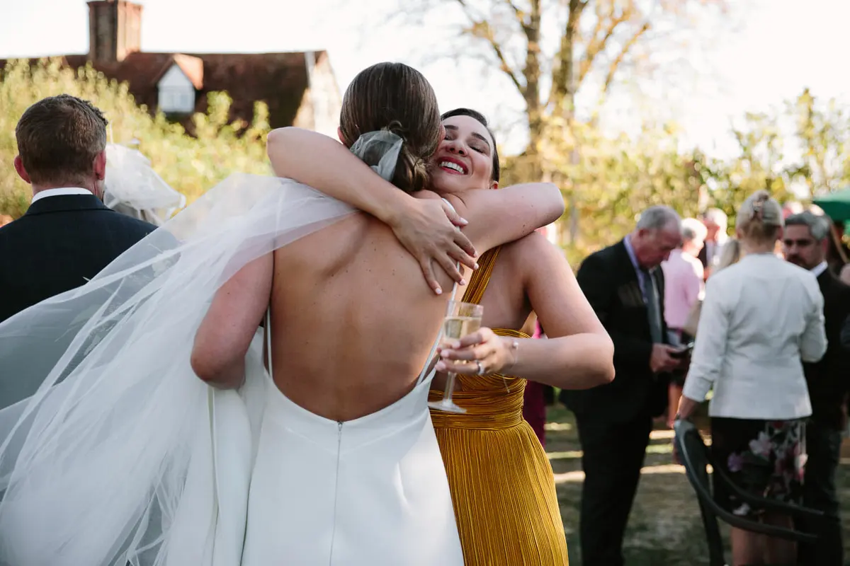 Bride in a veil hugging a laughing guest in a mustard dress at the outdoor drinks reception