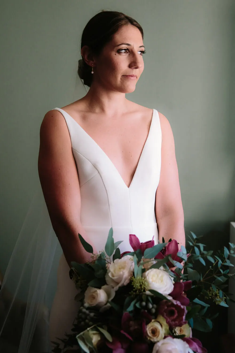 Bride in a V-neck dress and veil holding a burgundy and white bouquet by a window
