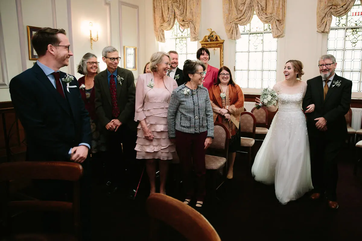 Bride entering on her father's arm as family stand and smile in a room with tall windows