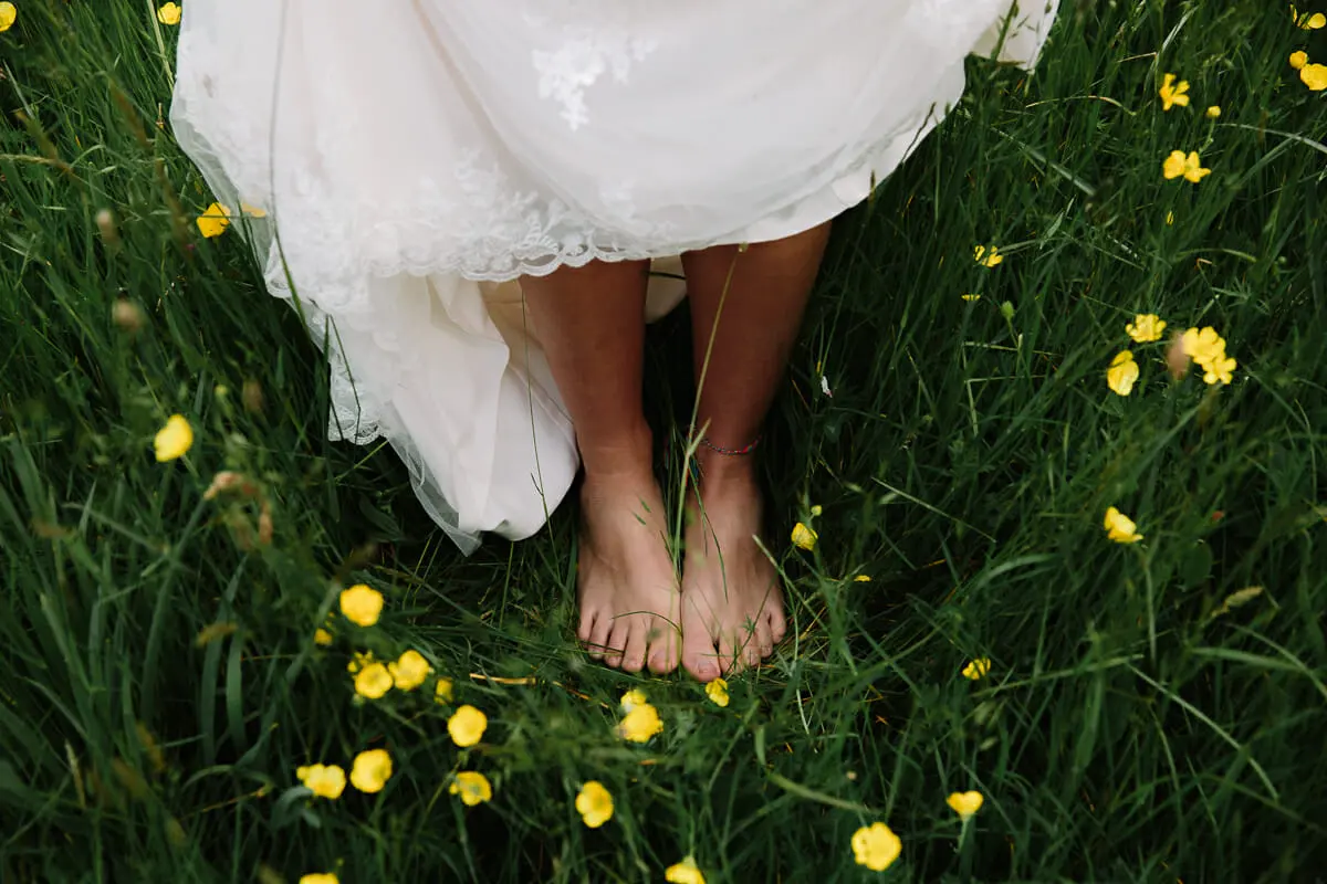 Bride's bare feet in long grass dotted with buttercups beneath the hem of her dress