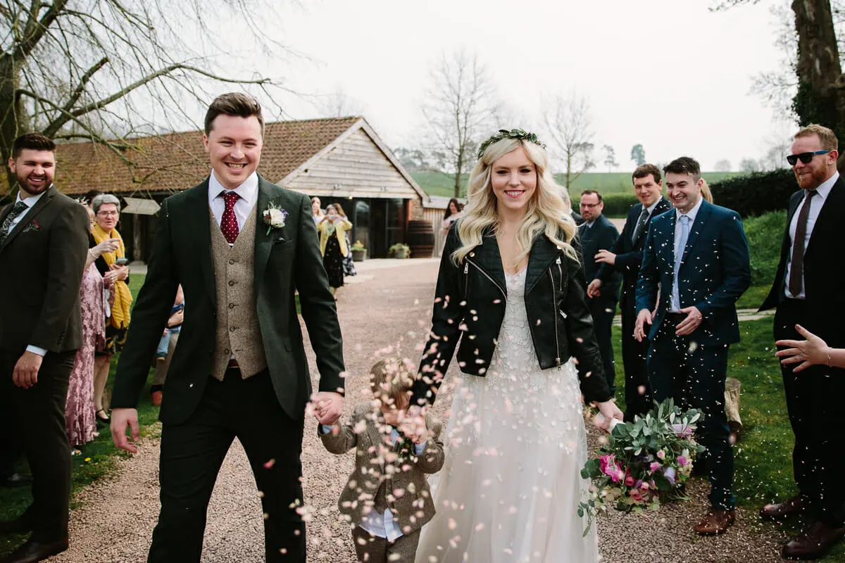 Bride in a leather jacket and groom walking hand in hand with a page boy as guests throw confetti