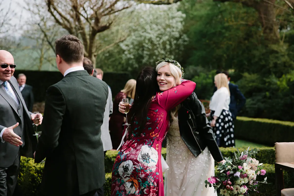 Bride in a leather jacket hugging a guest in a red floral dress during the garden drinks reception