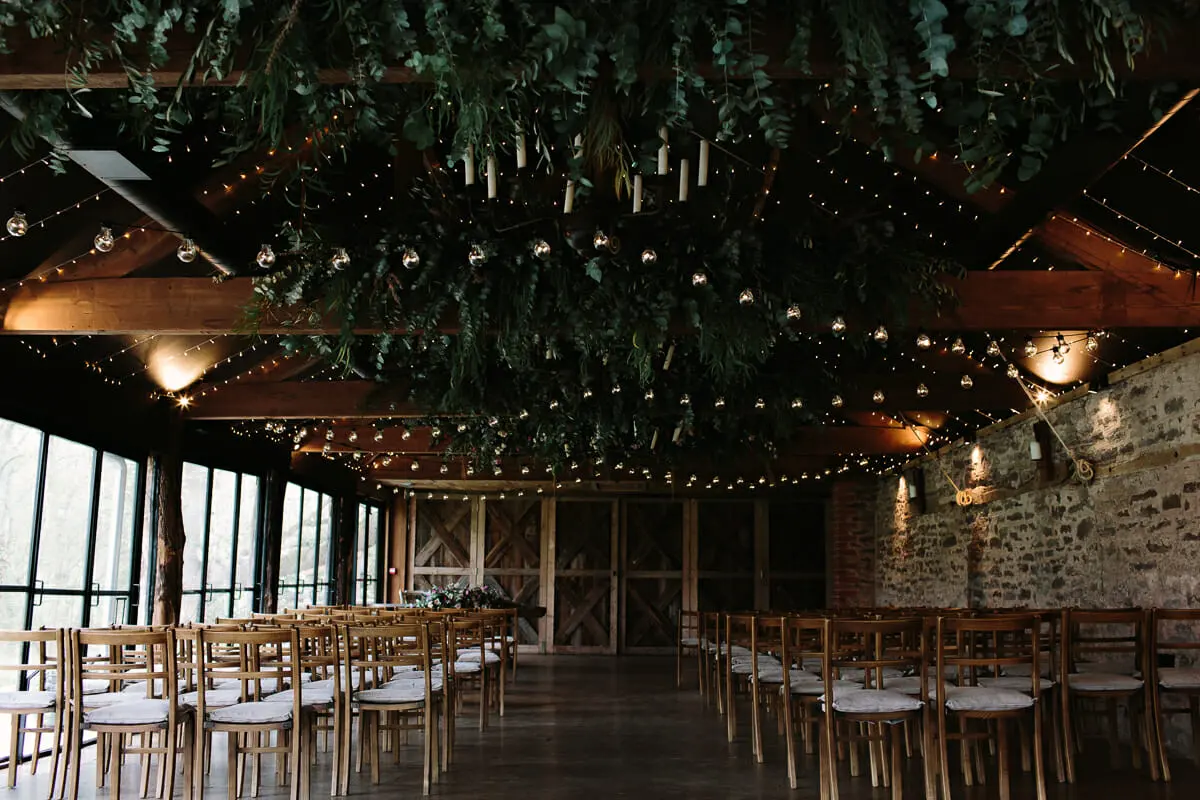 Empty ceremony room with rows of wooden chairs under a ceiling of hanging greenery and festoon lights
