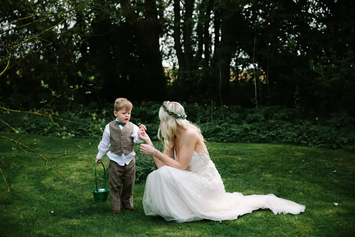 Bride kneeling on the lawn handing a flower to a page boy holding a small green basket