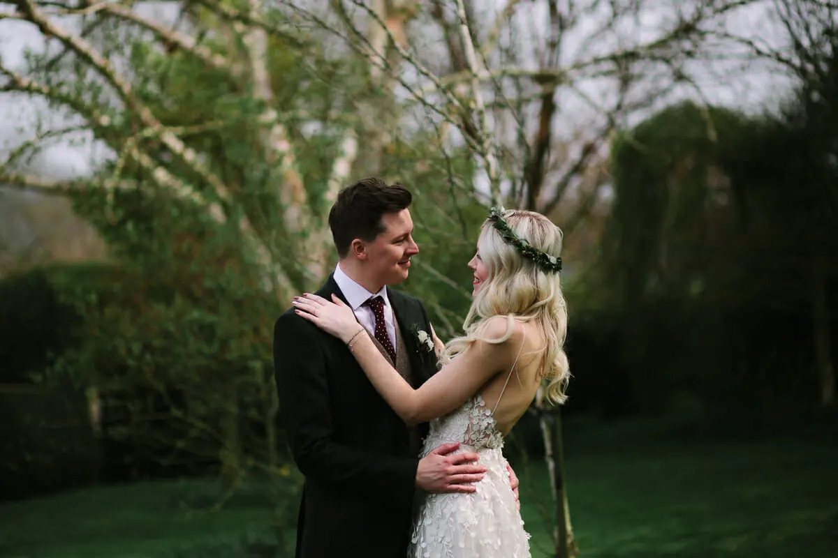 Bride in a flower crown with her hand on the groom's shoulder as they gaze at each other under birch trees