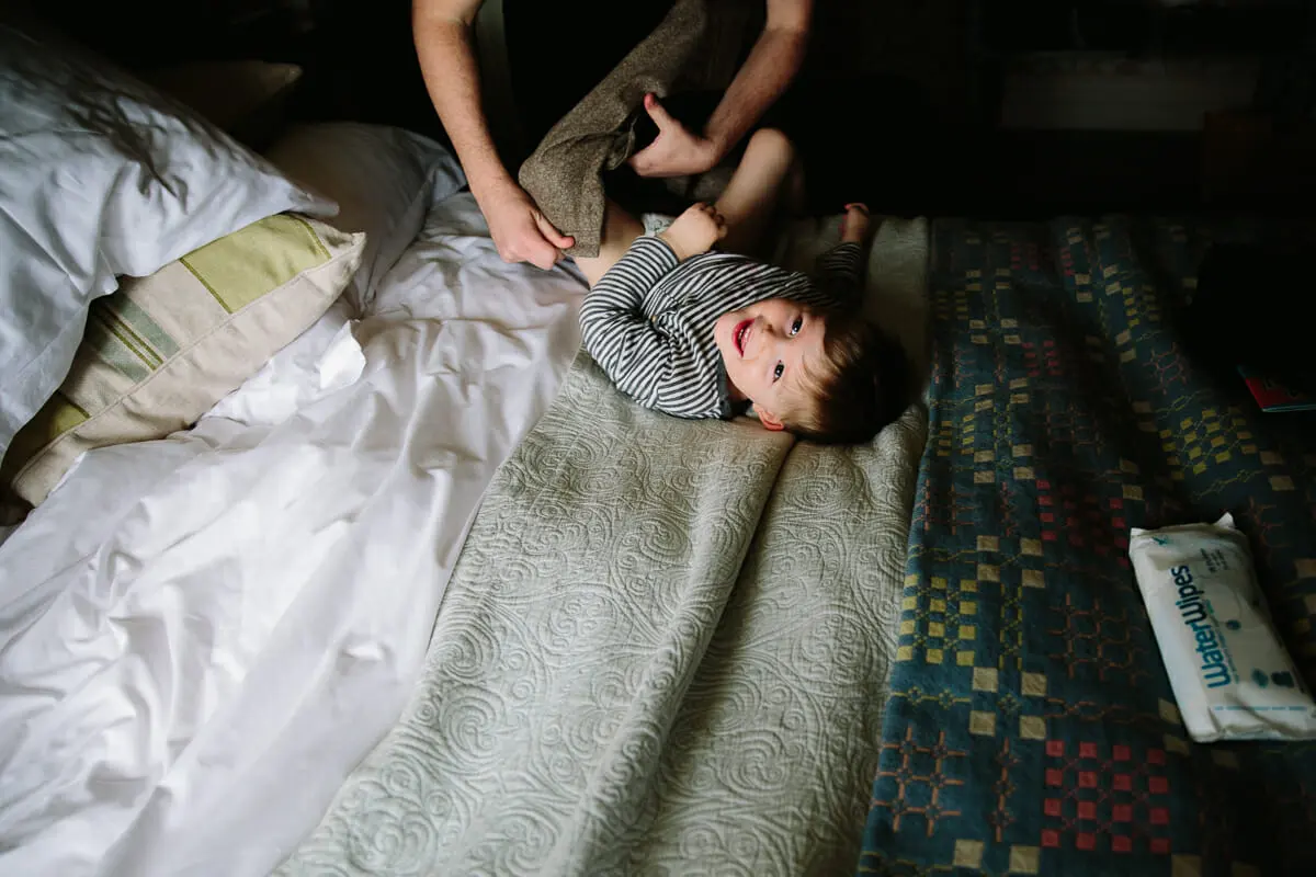 Young boy in a striped top lying on a bed laughing as an adult dresses him on the wedding morning