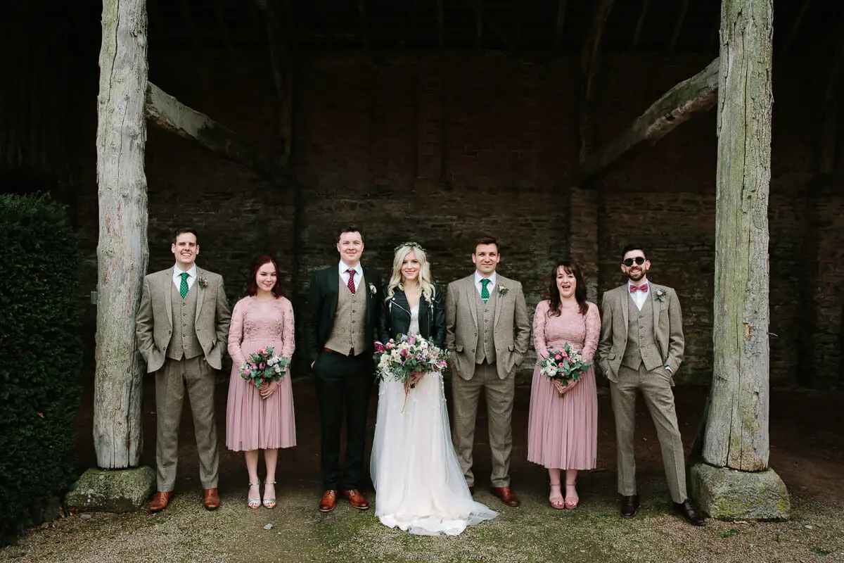 Bride and groom with bridesmaids in pink and groomsmen in tweed suits inside an old timber barn