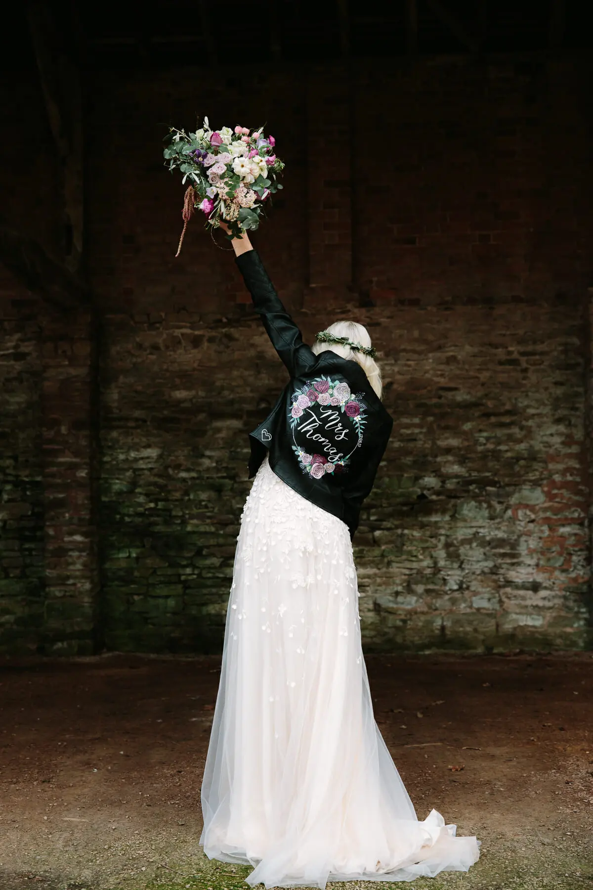 Bride seen from behind in a painted leather jacket, raising her bouquet in the air in an old barn