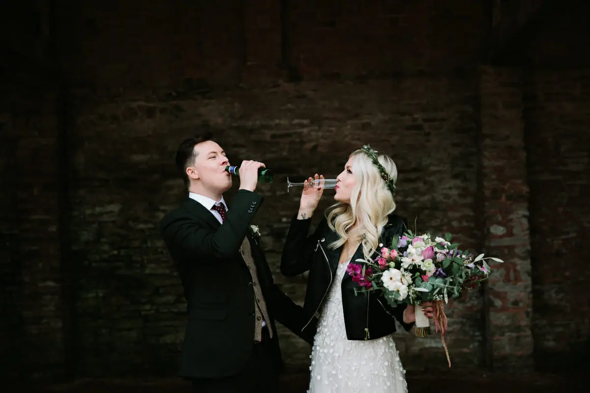 Groom drinking from a beer bottle and bride from a champagne flute against a dark brick wall