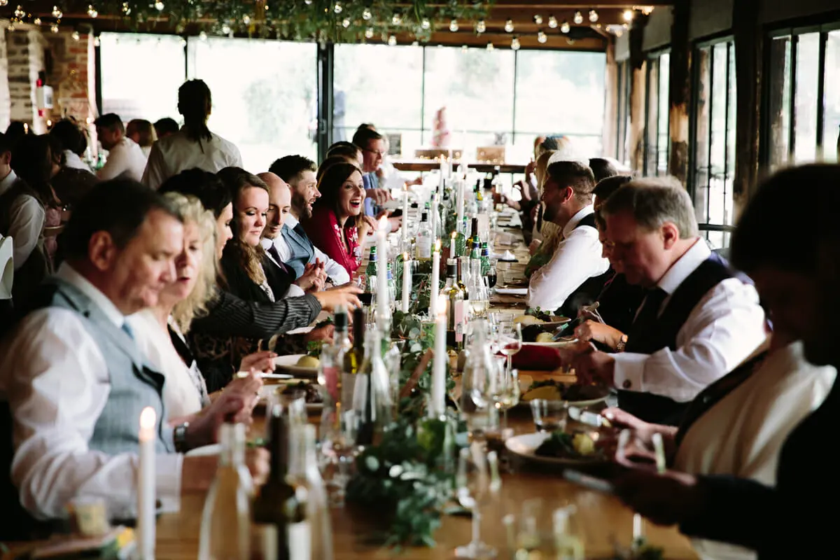 Guests eating the wedding breakfast at long candlelit tables under festoon lights in a barn