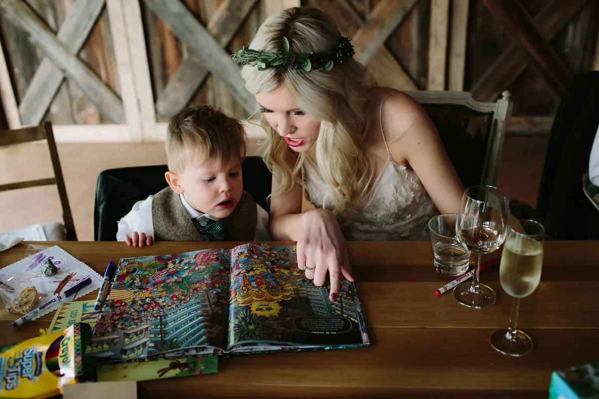 Bride in a flower crown reading a picture book with a page boy at the reception table