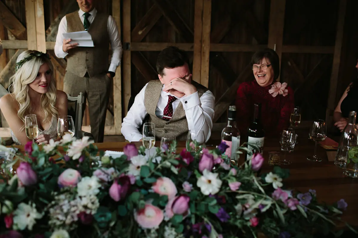 Groom covering his face, laughing, during a speech as the bride smiles beside him