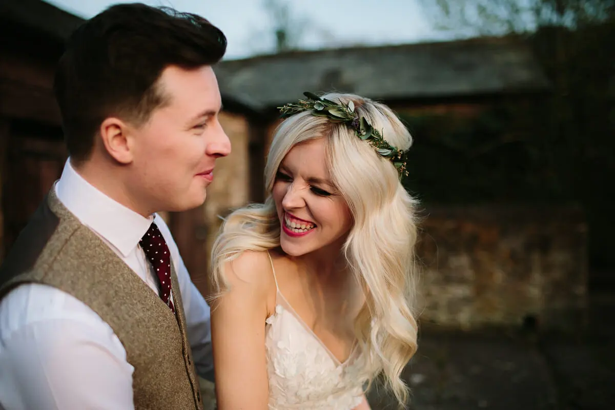 Bride in a leafy crown laughing with the groom in a tweed waistcoat outside a rustic barn
