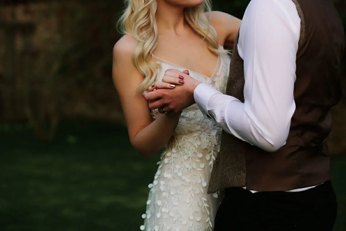 Groom holding the bride from behind, her hand on his, in a lace wedding dress outdoors
