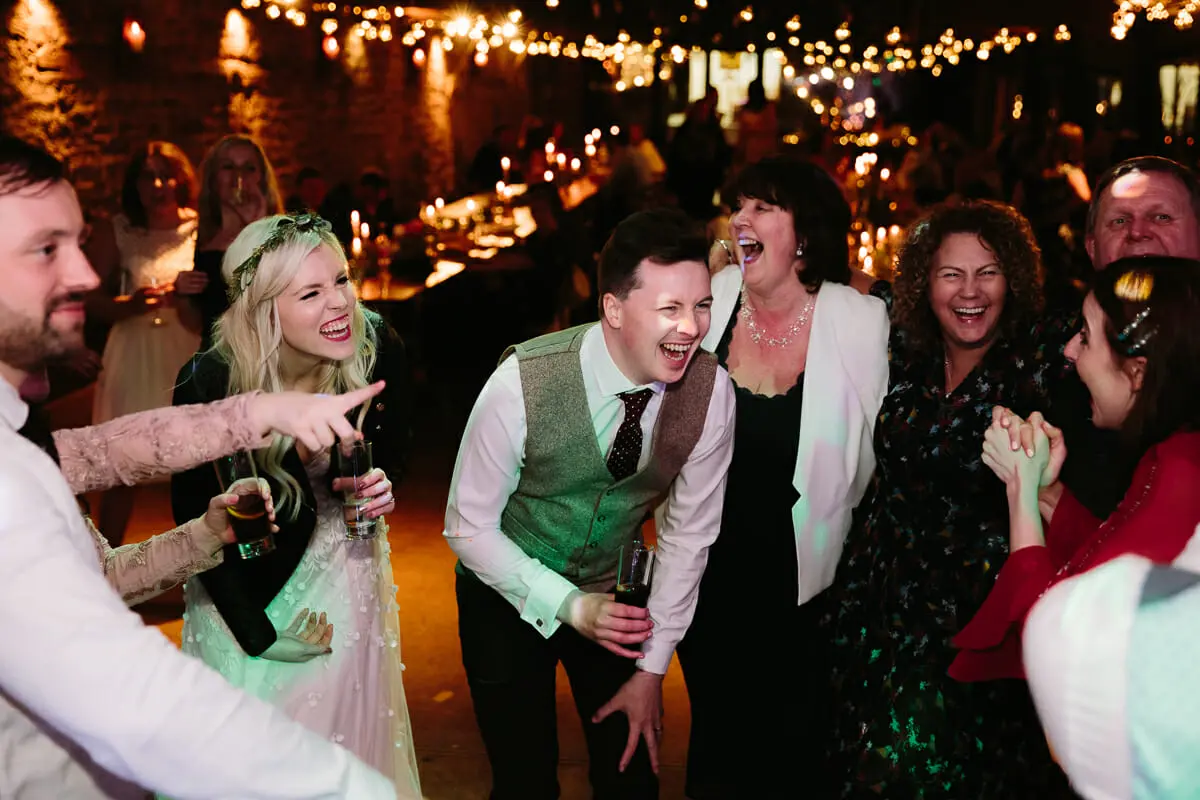 Bride and groom laughing with guests on a dance floor under strings of fairy lights