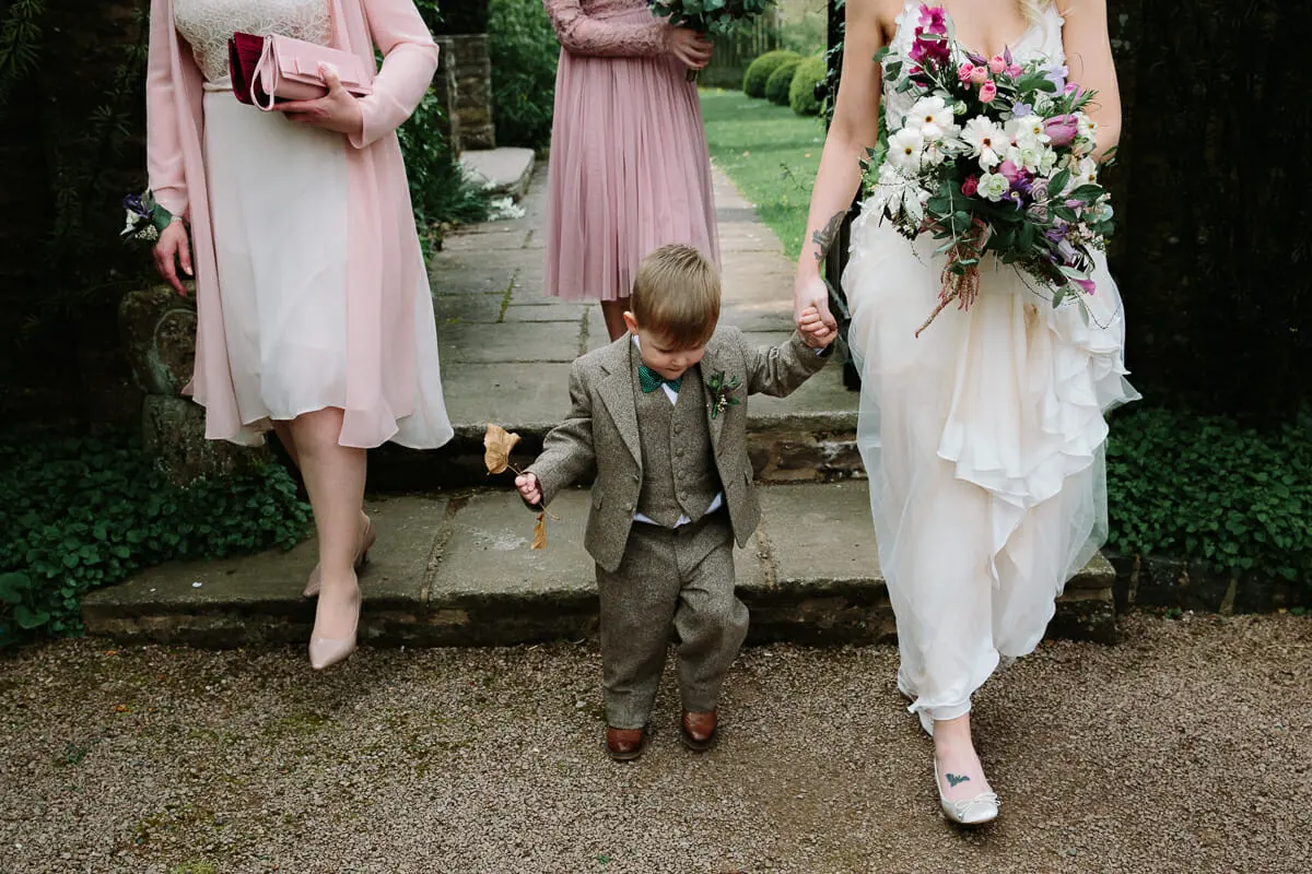 Page boy in a tweed suit holding a leaf as he walks hand in hand with the bride, bridesmaids behind