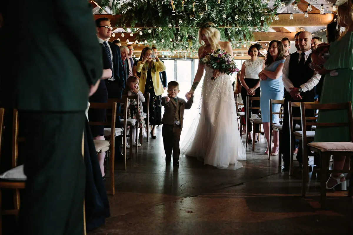 Bride walking down the aisle hand in hand with a page boy as guests watch under hanging greenery