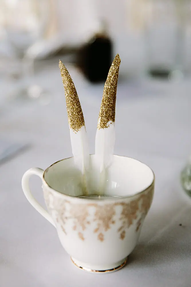 Two white feathers with gold glitter tips standing in a gold-patterned china teacup on a table