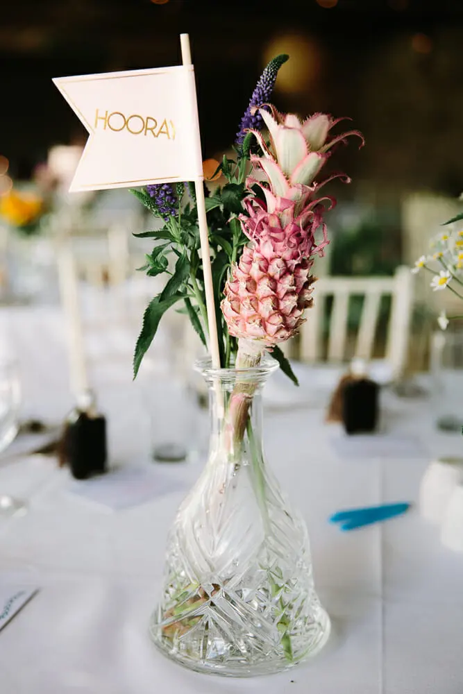 Small pink pineapple and purple flowers in a cut glass decanter with a Hooray flag on a reception table