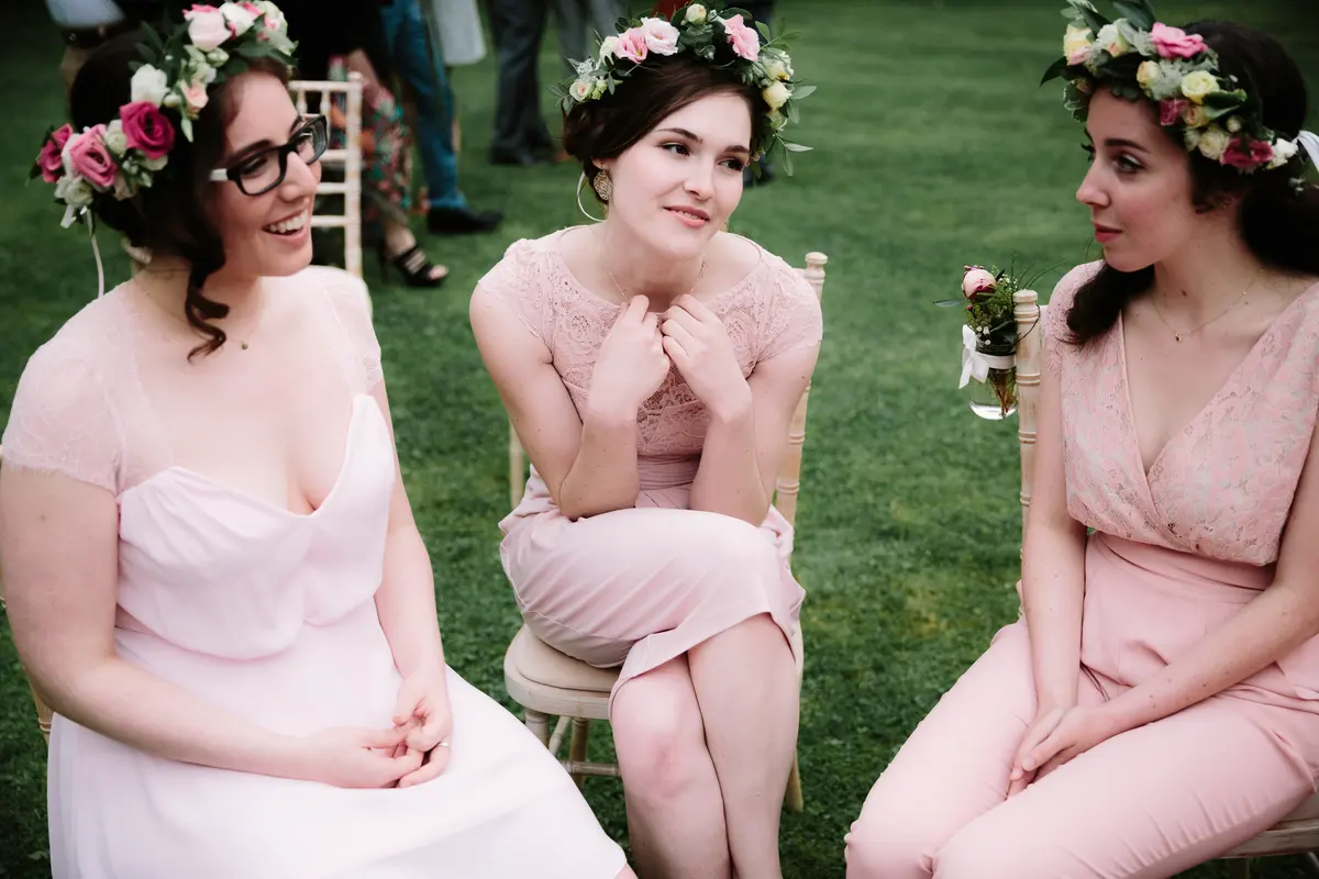 Three bridesmaids in pink with flower crowns chatting on chairs on a lawn