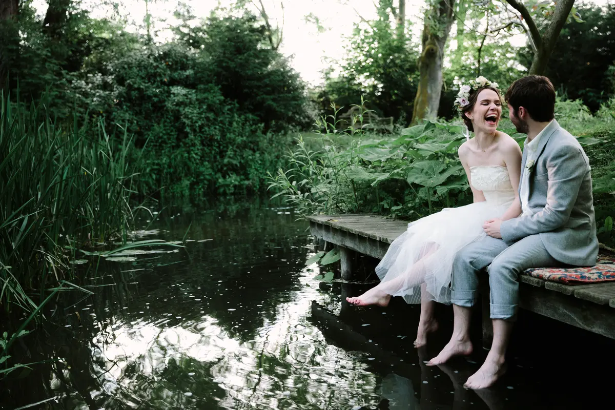 Barefoot bride and groom laughing on a jetty with their feet dangling in the pond