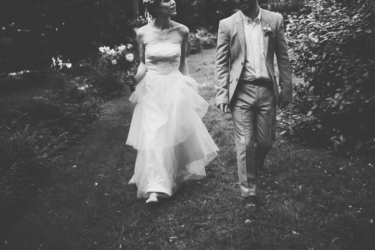 Black and white shot of the bride and groom walking along a grassy woodland path