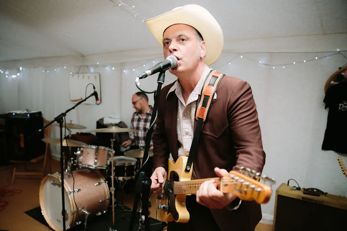 Singer in a cowboy hat playing electric guitar with a drummer behind in a fairy-lit marquee