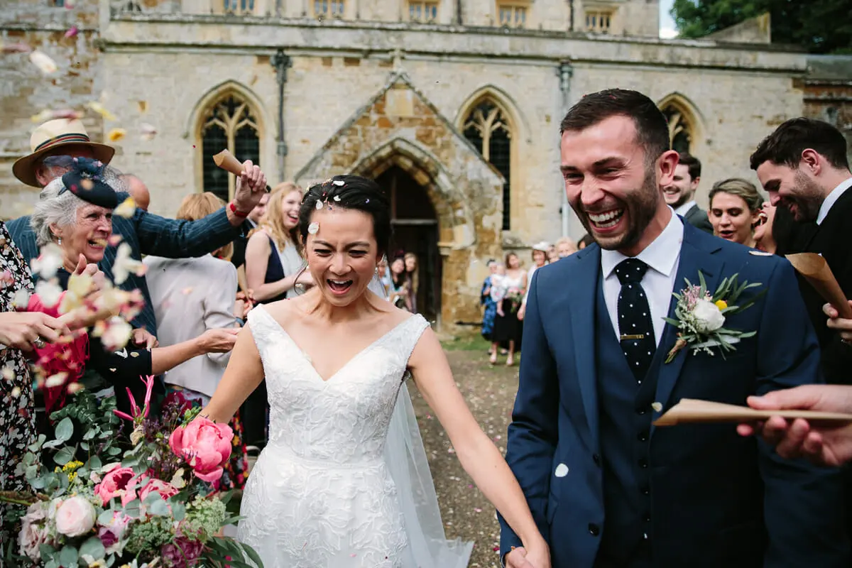 Bride and groom laughing hand in hand as confetti flies outside the church