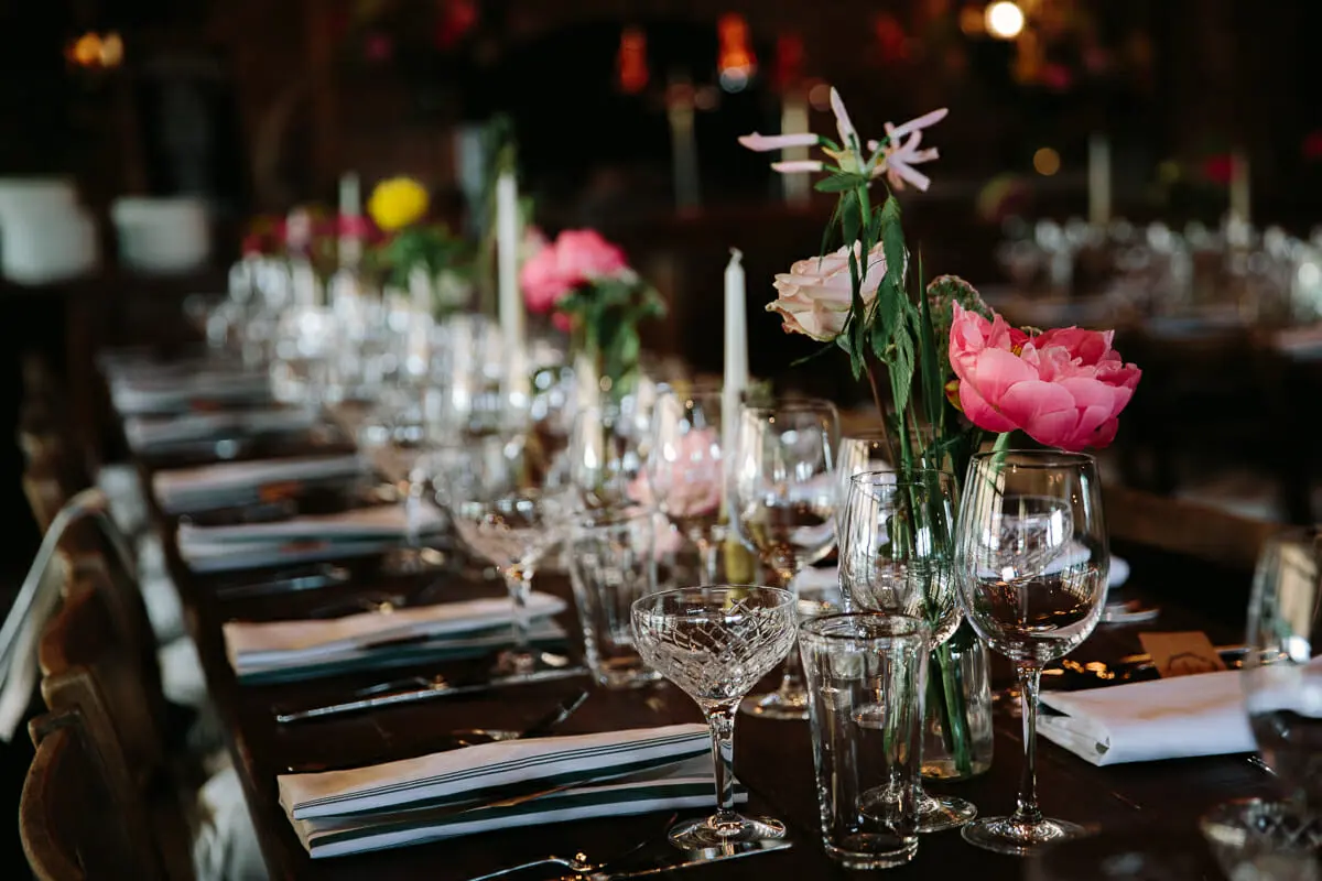 Long table laid with glassware, candles and vases of pink peonies in the barn