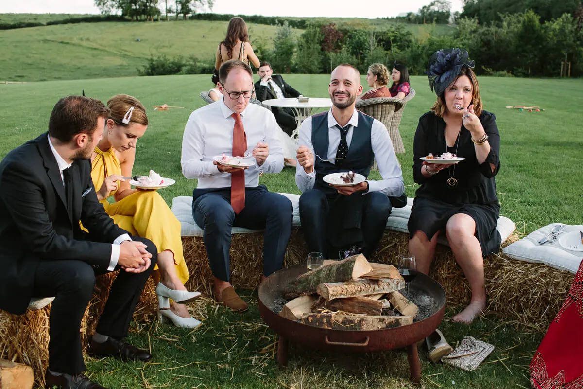 Guests eating cake on hay bales around a fire pit on the lawn at Soho Farmhouse